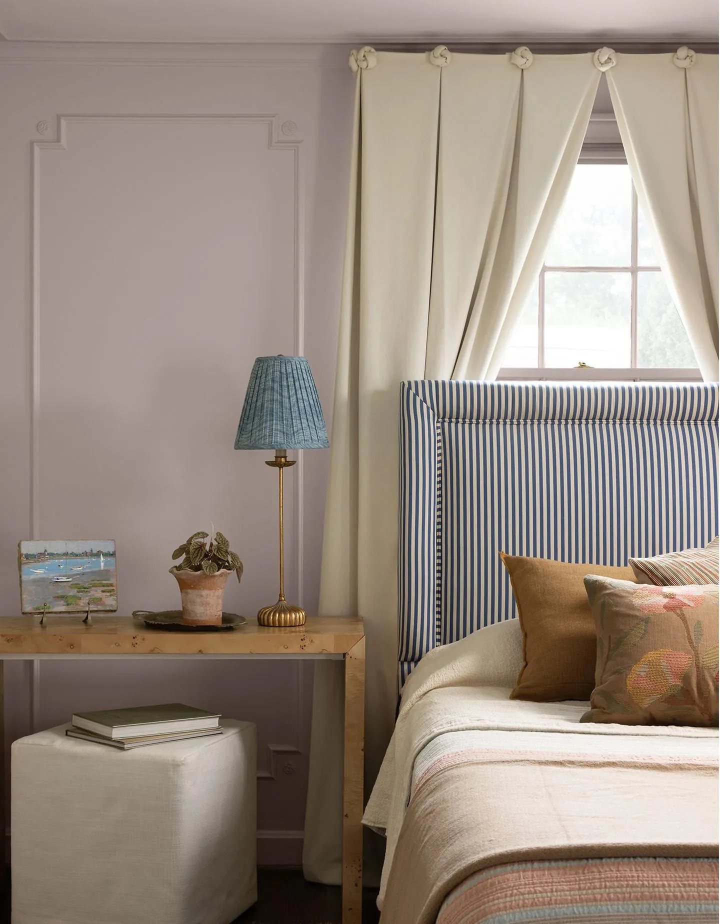 A bedroom painted in Wild Aster with a striped upholstered headboard, white linens, and a wood desk